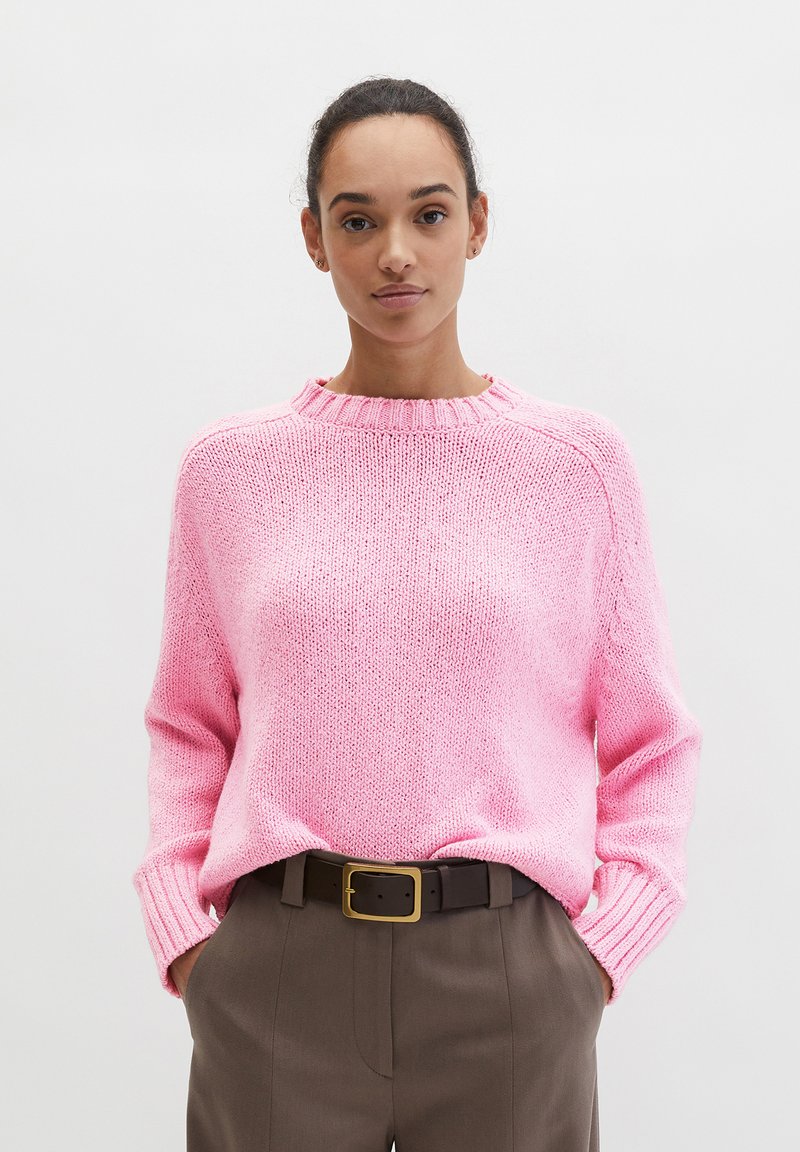 Femme portant un pull en maille rose rentré dans un pantalon marron taille haute avec une ceinture noire, debout devant un fond clair uni.