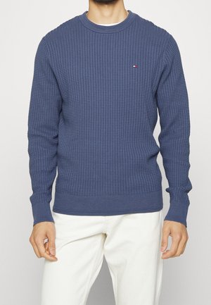 Blaues Sweatshirt aus Waffelstrick mit langen Ärmeln, geripptem Halsausschnitt und Bündchen, mit kleinem Logo auf der Brust. Kombiniert mit weißen Hosen.