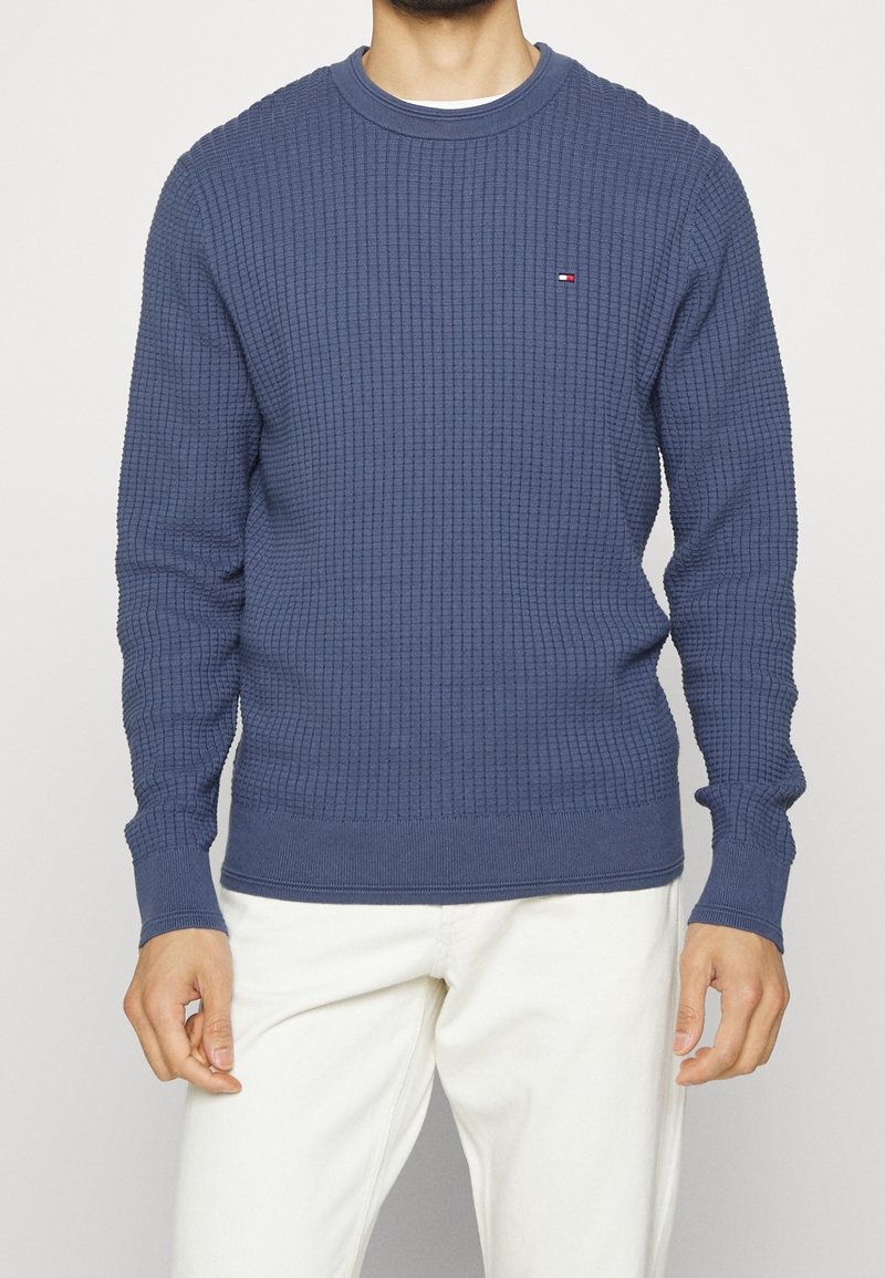 Blaues Sweatshirt aus Waffelstrick mit langen Ärmeln, geripptem Halsausschnitt und Bündchen, mit kleinem Logo auf der Brust. Kombiniert mit weißen Hosen.