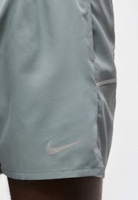 Γκρι αθλητικό σορτς Nike με ανακλαστικό λογότυπο swoosh, φορεμένα στο πάνω μέρος του μηρού ενός ατόμου με απλό φόντο.