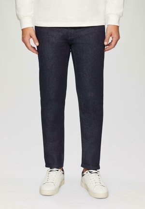 Donkerblauwe denimjeans met een slim fit, voorzien van een gladde textuur en subtiele stiksels. Gecombineerd met witte sneakers voor contrast.