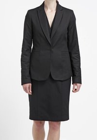 Svart skräddarsydd kavaj med en enkel knappstängning och två sidofickor, i kombination med en matchande knälång pennkjol. Slät textur.