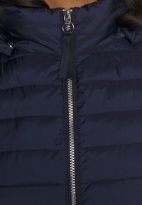 Tamsiai mėlynas pūkinis švarkas su užtrauktuku, horizontalios quilted tekstūros ir aukštu apykakle. Užtrauktukas turi sidabrinį apdailą ir juodus akcentus.