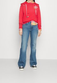 Červená mikina s kapucí a bílými stahovacími šňůrkami, s vyšitými detaily, vrstvená přes vícebarevnou károvanou košili, doplněná rozšířenými modrými džínami a černými teniskami.