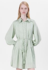 Hellgrünes langärmliges Kleid mit durchgeknöpftem Vorderteil, strukturiertem Stoff, Rüschendetails an den Schultern, tailliertem Bund und Quastenbändern.