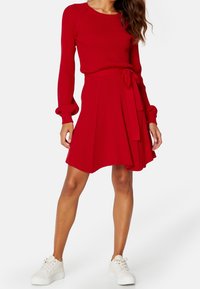 Robe tricotée rouge à côtes avec des manches longues et une taille nouée, dotée d'une jupe évasée. Associée à des baskets blanches épaisses.