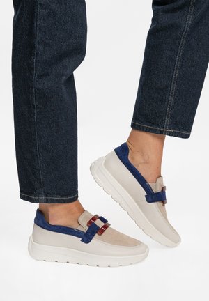 Person trägt beige und marineblaue Slip-On-Sneaker mit kastanienbraunen Schnallenakzenten und dunkelblaue Jeans.