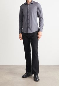 Camisa gris de botones con cuello, mangas largas y un logo sutil, combinada con pantalones negros acampanados y zapatos negros con cordones.