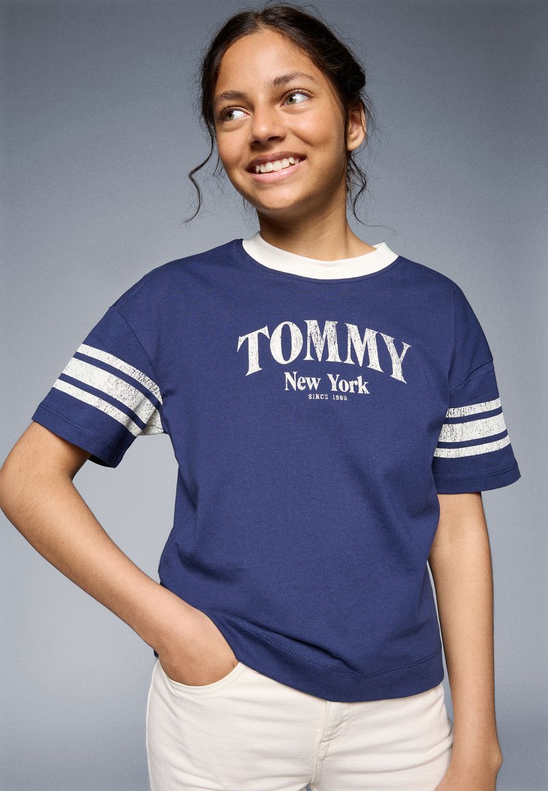 Mörkblå bomullströja med vit ringerkrage, "TOMMY New York" tryck och metallicrandiga ärmar. Kort, avslappnad passform.