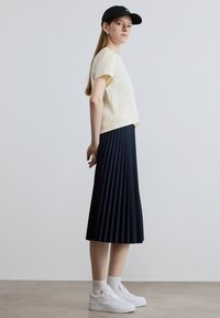 Ανοιχτό κίτρινο τοπ με κοντά μανίκια, ναυτικό πλισέ φουστάνι, λευκά αθλητικά παπούτσια και μαύρο καπέλο. Ομαλή υφή, κλασική σιλουέτα και λιτός σχεδιασμός.