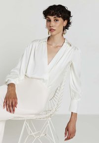 Blusa bianca in satin con scollatura a V profonda, maniche lunghe a sbuffo e una texture liscia, abbinata a pantaloni chiari e una sedia di metallo.