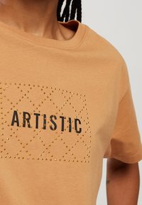 Beżowa koszulka bawełniana z napisem "ARTISTIC" w czarnym nadruku, ozdobiona złotymi ćwiekami ułożonymi w diamentowy wzór.