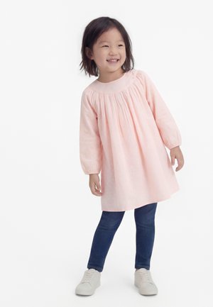 Smilende ung pige iført en lyserød kjole, mørkeblå leggings og hvide sneakers, stående mod en hvid baggrund.