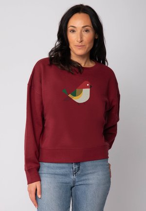 Vrouw in een bordeaux sweatshirt met een geometrisch vogeldesign op de borst en lichtblauwe jeans, staand tegen een effen lichte achtergrond.
