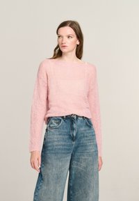 Helles rosa gestricktes Sweatshirt mit weitem Halsausschnitt und langen Ärmeln, kombiniert mit lockeren blauen Jeans. Strukturierter Stoff sichtbar.
