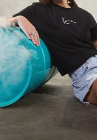 Svart bomull T-shirt med vit skrivlogga, ihopparat med ljusblå denimshorts. Lutar mot en turkos tunna med en strukturerad yta.