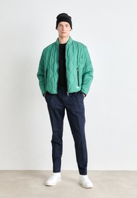 Veste bomber verte matelassée avec col haut, zips sur les côtés, pull noir, pantalon bleu marine et baskets blanches. Tissu texturé avec un motif en losanges.
