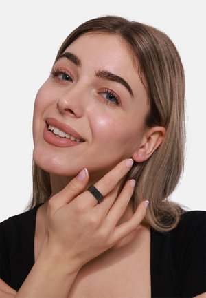 Bague en métal noir avec un design rainuré, portée à l'index, sur un fond neutre. Le modèle a la peau claire et de longs cheveux.
