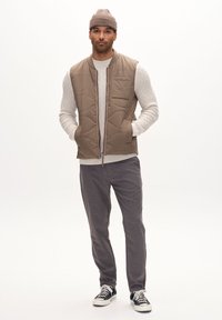 Beige padded vest with a zip front, textured light knit sweater underneath, gray patterned trousers, and black-and-white sneakers.