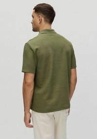 Koszulka polo z krótkim rękawem w oliwkowym kolorze z prążkowanej tkaniny, z klasycznym kołnierzykiem i bocznymi rozcięciami, w zestawie z jasnymi spodniami.