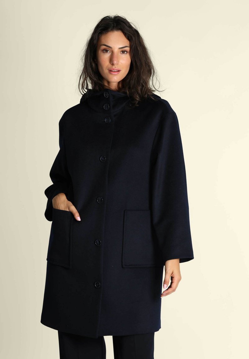 Cappotto in lana blu navy con un alto collo, sette grandi bottoni e due tasche frontali. Tessuto liscio e vestibilità rilassata con maniche a tre quarti.