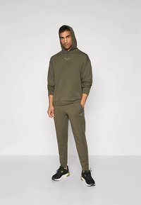 Olivovo zelená mikina s kapucňou a zodpovedajúce tepláky vyrobené z mäkkého materiálu, s predným vreckom, sťahovacími šnúrkami a čiernymi športovými topánkami.