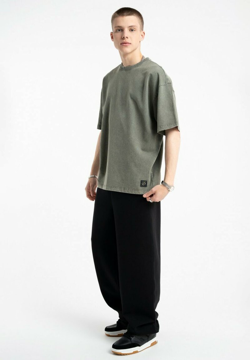 Olive zielony oversize'owy t-shirt bawełniany z okrągłym dekoltem i krótkimi rękawami, zestawiony z luźnymi czarnymi spodniami i czarnymi trampkami.