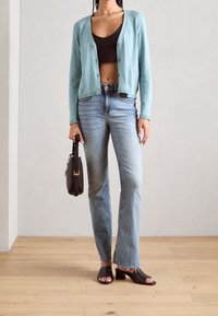 Un cardigan bleu clair sur un crop top noir, associé à un jean droit de teinte moyenne et des talons ouverts noirs. Tenant un petit sac noir.