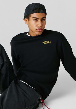 Young man wearing black Red Bull Racing sweatshirt and black cap, sitting and leaning back against a plain light background.