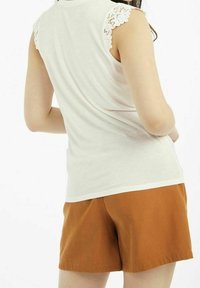 Bílý bezrukávový top s krajkovými detaily na ramenech, spárovaný s hnědými bavlněnými šortkami. Jednoduchý design s jemnou texturou a volným střihem.