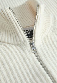 Maglione a coste color crema con colletto alto e zip argentata. Presenta un tessuto testurizzato e un'etichetta del marchio visibile vicino allo scollo.