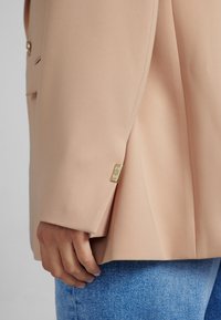 Béžový blazer vyrobený z hladkého materiálu, se zlatým logem na rukávu. Nosený s světle modrými džínami.