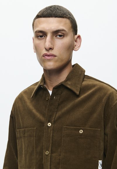Brown corduroy shirt with a button-up front, two chest pockets, and a spread collar. Vertical ribbed texture with a white logo tag at the hem.