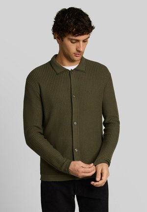 Man met krullend haar die een donkergroene, gestructureerde cardigan met knopen draagt en de mouwboord aanpast tegen een effen grijze achtergrond.