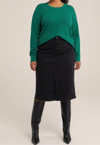 Grön texturerad stickad kofta med knappstängning, kombinerad med en svart jeanskjol till knäna med en slits framtill, bärs med svarta knähöga stövlar.