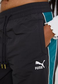 Black athletic pants with teal side panels, elastic waistband, and logo detail. Made from lightweight material, featuring a side pocket.