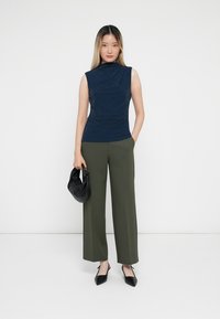 Top sin mangas de color azul marino con tejido texturizado, combinado con pantalones de pernera ancha en color verde oliva. La modelo sostiene un bolso negro y lleva zapatos negros de punta.