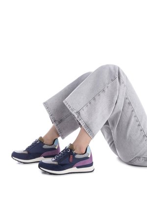 Zapatillas azul marino con acentos de gamuza y malla, con detalles en morado y azul claro, suela blanca y diseño con cordones, colocadas sobre una figura sentada.