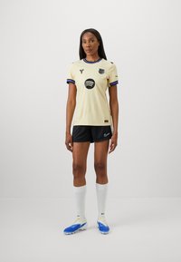 Ljusgul fotbollströja med blå detaljer, med en central logotyp och svarta inslag. Kombination med svarta shorts och vita strumpor.