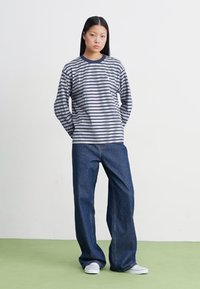Camisa de manga longa às riscas com linhas horizontais em azul-marinho e branco, combinada com jeans soltos de cor azul-escuro e ténis brancos, de pé sobre um chão verde.