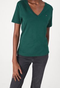 Grön kortärmad V-ringad t-shirt i bomull som har en liten logotyp på vänstra bröstet. Matchad med mörkgrå jeans.