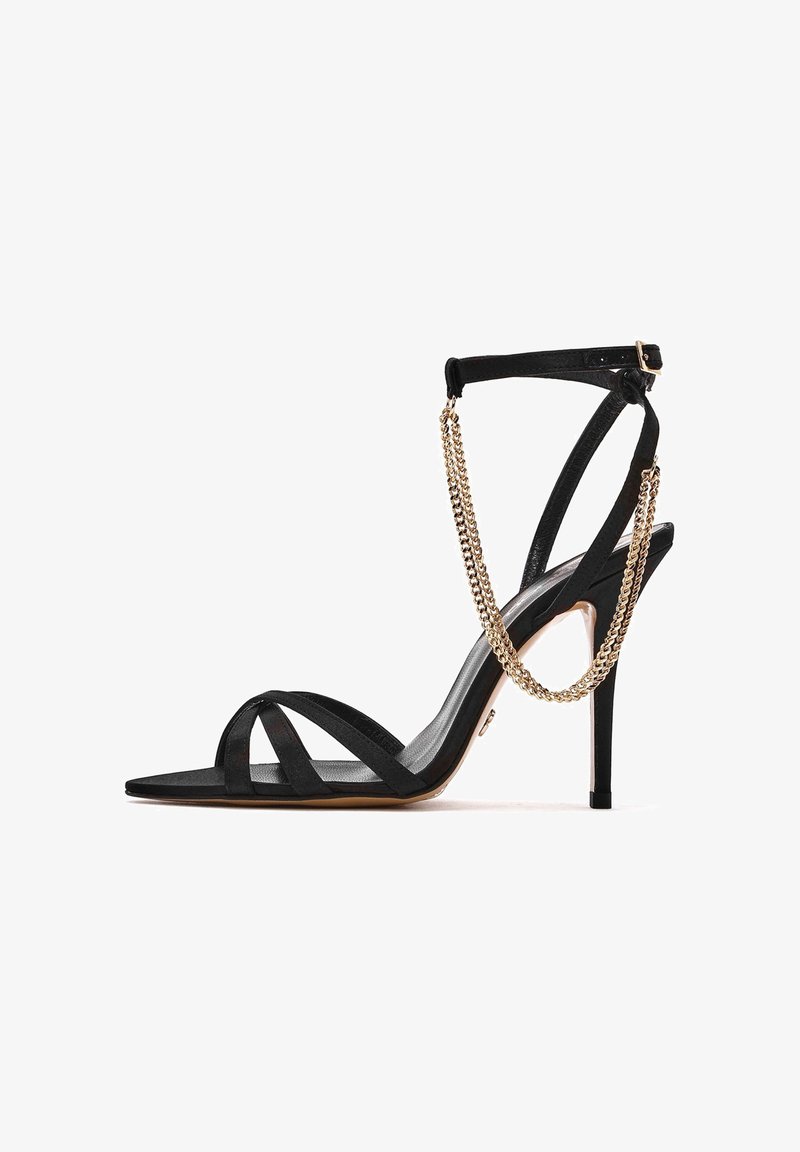Zwarte stiletto sandaal met meerdere dunne banden, een spitse neus, een suède textuur en een gouden kettingaccent op de enkelband.
