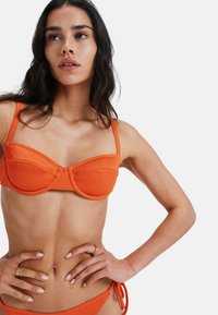 Top de bikini portocalie cu nervuri, cu suport subțire, având bretele late și cusături curbate. Textura netedă evidențiază culoarea sa îndrăzneață și designul structurat.