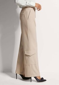 Beige wijde broek met een trekkoord in de taille, zijzakken en details van cargozakken, gecombineerd met zwarte pumps met een spitse neus. Glad stoftextuur.