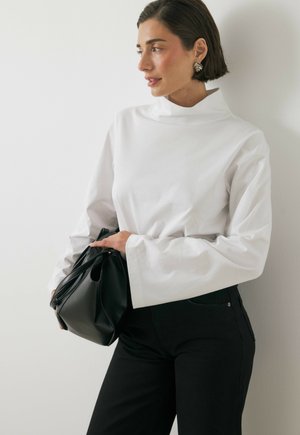 Woman with short dark hair wearing white long-sleeve top and black pants, holding black handbag, looking to the right against a light wall.