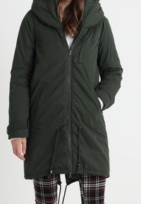 Žena v tmavozelené parce s kapucí a zipem, stojící s jednou rukou v kapse a v kostkovaných kalhotách.