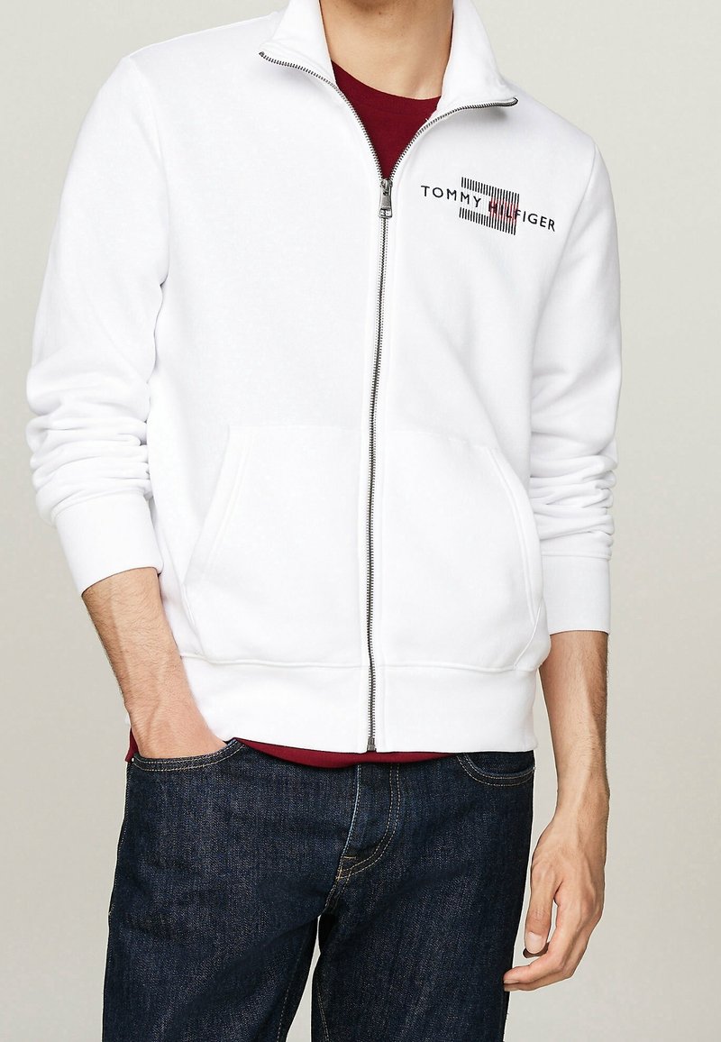 Sudadera blanca con cremallera y mangas largas, que presenta un logo negro y un diseño a rayas en el pecho. Tejido suave, corte estándar y bolsillos delanteros.