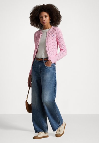 Woman with curly hair wearing a pink cable-knit cardigan, white shirt, wide-leg jeans, beige sneakers, holding a brown handbag.