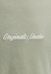 Tissu vert pâle avec texte brodé en blanc indiquant "Originals Studio" en police cursive.