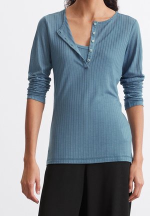 Frau trägt ein blaues, geripptes Langarmshirt mit teilweise geöffneten Druckknöpfen, kombiniert mit schwarzer Hose, vor einem schlichten weißen Hintergrund.